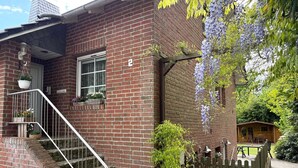 A brick house with a porch, a wooden fence, and a garden with purple flowers. - Kirsten 5 - Cozy vacation apartment close to the beach for up to 4 people - House (Cuxhaven)