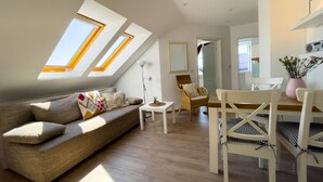 A modern living room with a sofa, dining table, and chairs. There are skylights on the ceiling, a lamp, and a vase with flowers. - Kirsten 5 - Cozy vacation apartment close to the beach for up to 4 people - House (Cuxhaven)