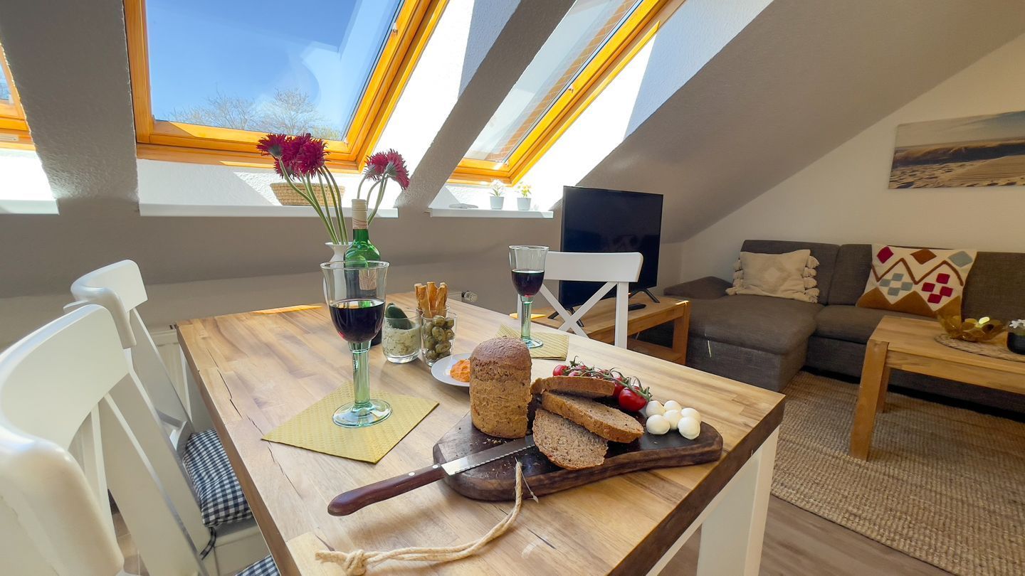 Ein Essbereich mit Holztisch, Messer, Brot und einer Weinflasche. Ein Fenster mit Blick zum Himmel und ein Gemälde an der Wand.