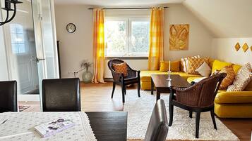 Ein Wohnzimmer mit gelber Couch, einem Esstisch mit Stühlen, einem Korbstuhl, einem weißen Teppich, einer Uhr und einem Fenster mit Vorhängen.