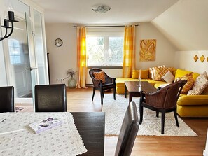 Un salon avec un canapé jaune, une table à manger avec des chaises, un fauteuil en osier, un tapis blanc, une horloge et une fenêtre avec des rideaux.