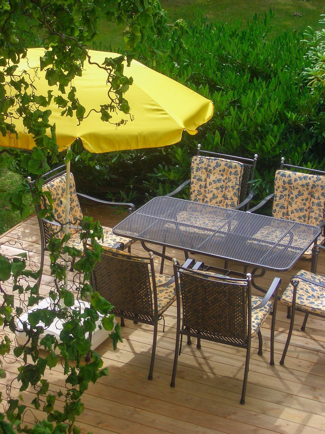 Eine Terrasse im Freien mit Tisch und Stühlen, einem gelben Sonnenschirm und Grünpflanzen im Hintergrund.
