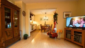 A living room with a TV, wooden furniture, a dining area, and a kitchen in the background.