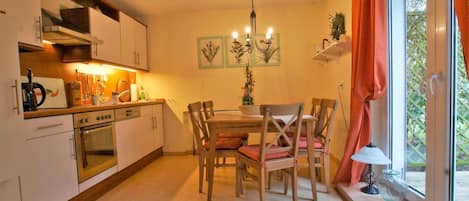 A kitchen with a dining table, chairs, and a chandelier. There is a window with curtains and a view of greenery outside.