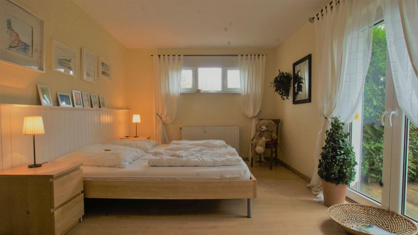 Ein Schlafzimmer mit einem Bett, Nachttischlampen, einem Sessel, einem Fenster mit Vorhängen und einer Zimmerpflanze.
