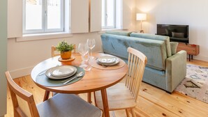 A dining area with a round table set for four, a sofa, a TV, and a window with blinds. - D&S -JB Apartments - B (Oporto)