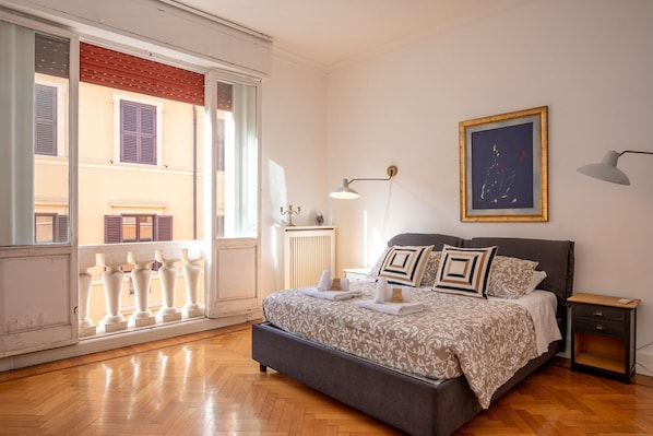 Ein Schlafzimmer mit Bett, Nachttisch, Lampe und einem Fenster mit Blick auf Gebäude.