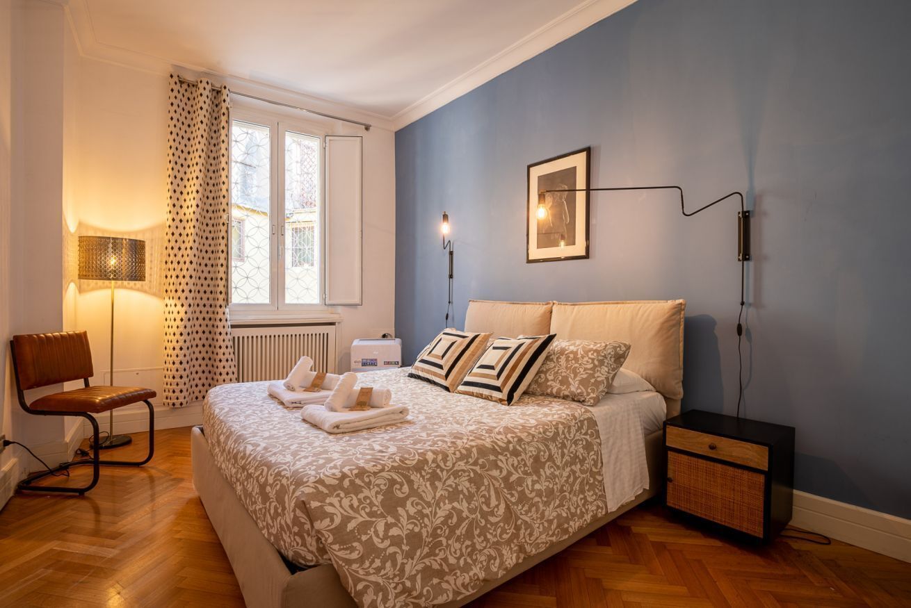 Een slaapkamer met een bed, een stoel, een nachtkastje, een lamp en een raam met gordijnen.