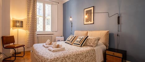 Een slaapkamer met een bed, een stoel, een nachtkastje, een lamp en een raam met gordijnen.