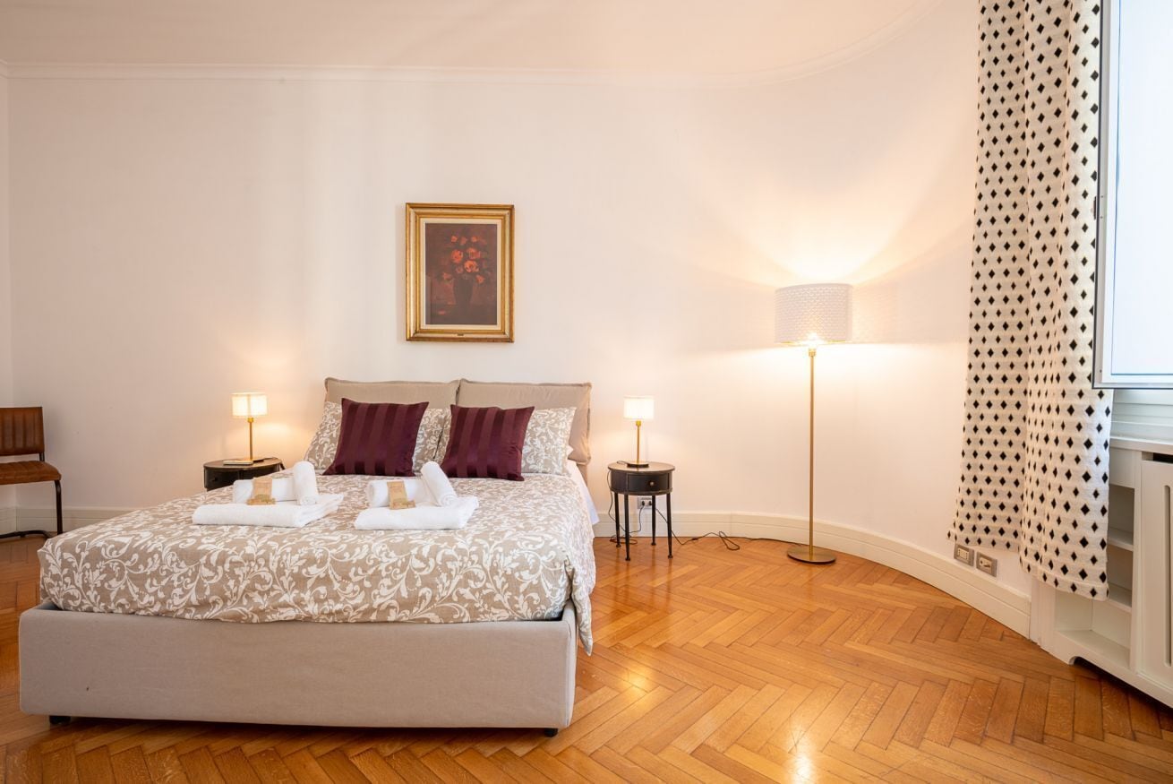 Un dormitorio ordenado con una cama, dos mesitas de noche, una lámpara de pie, un cuadro en la pared y una cortina estampada.