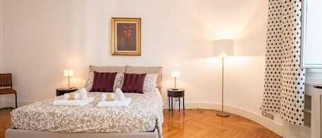 Un dormitorio ordenado con una cama, dos mesitas de noche, una lámpara de pie, un cuadro en la pared y una cortina estampada.