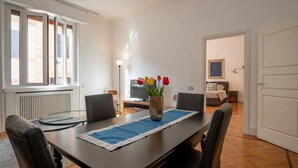 Spiseområde med bord, stole, en vase med blomster, en radiator, et fjernsyn og en seng i baggrunden.