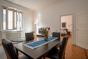 Zona pranzo con tavolo, sedie, un vaso con fiori, un termosifone, una televisione e un letto sullo sfondo.
