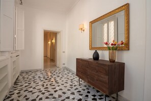 Un corridoio con pavimento piastrellato, un comò in legno, uno specchio e un vaso con fiori.
