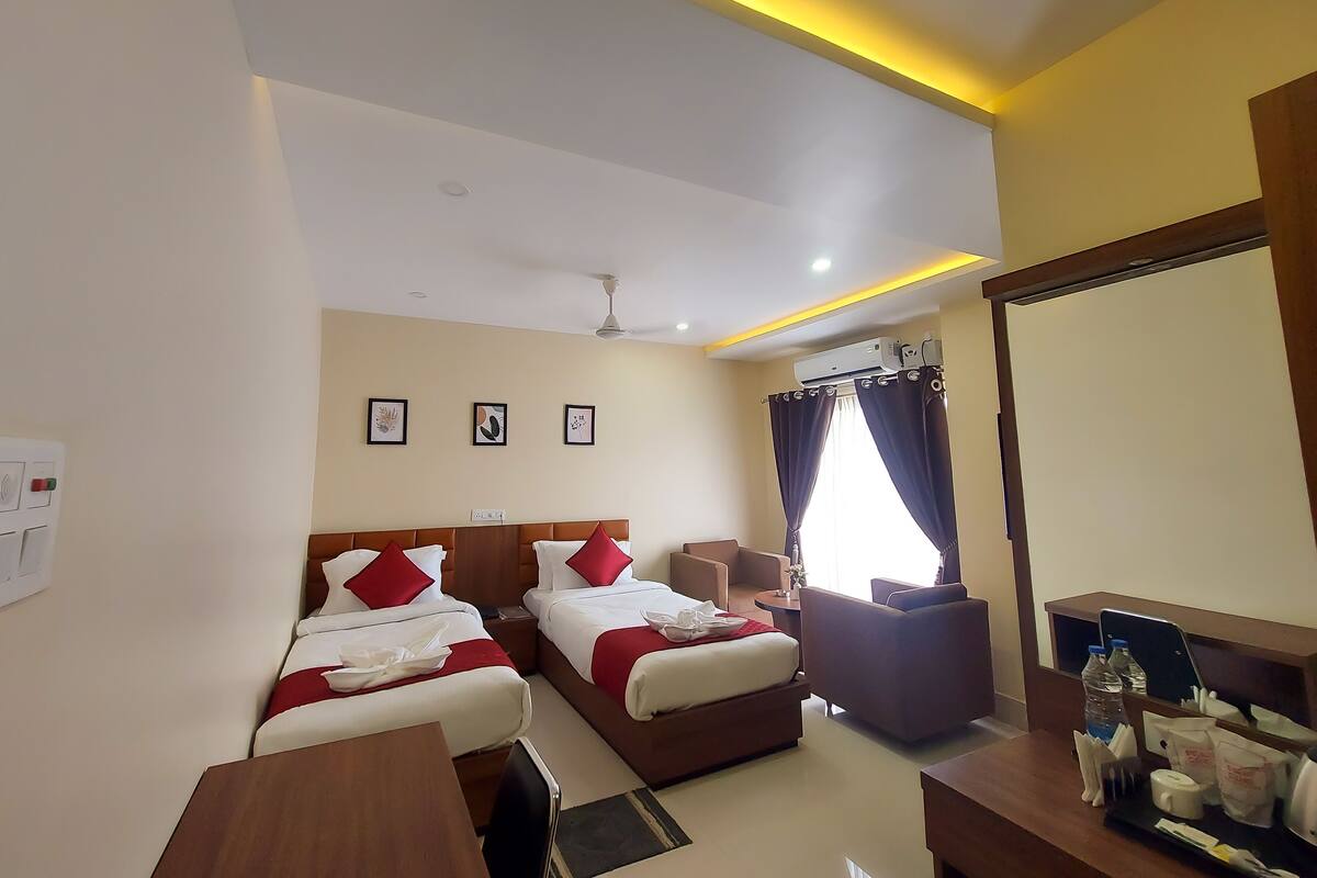 A hotel room with two beds, a desk, a chair, a small table, a mirror, and an air conditioning unit.