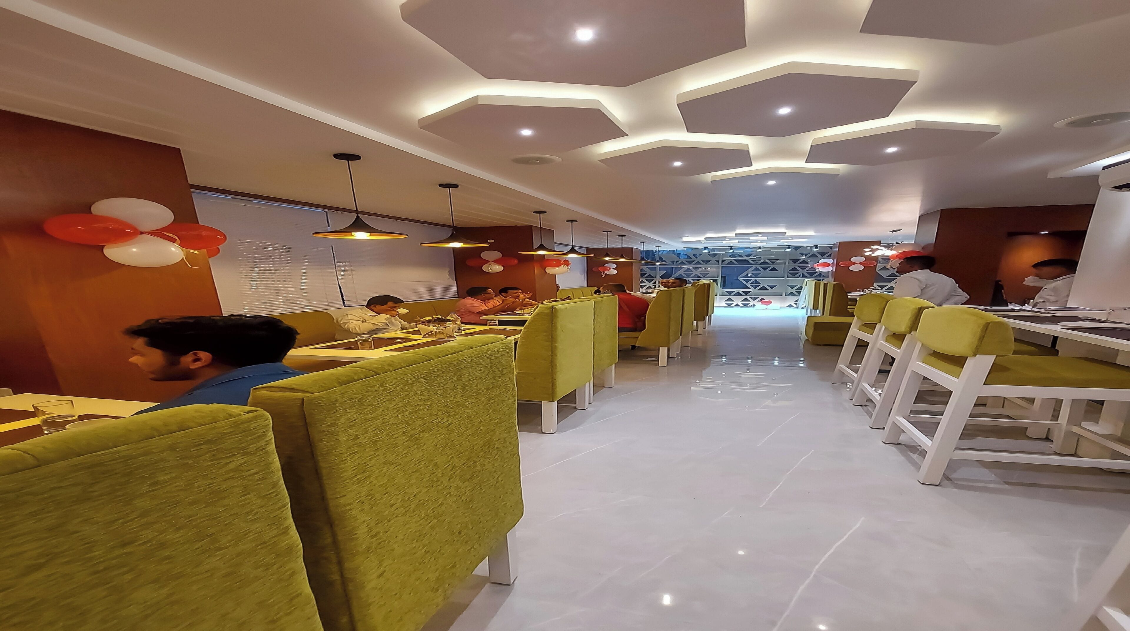 Un restaurante moderno con asientos tapizados en verde y mesas blancas, que presenta un diseño geométrico en el techo e iluminación ambiental.
