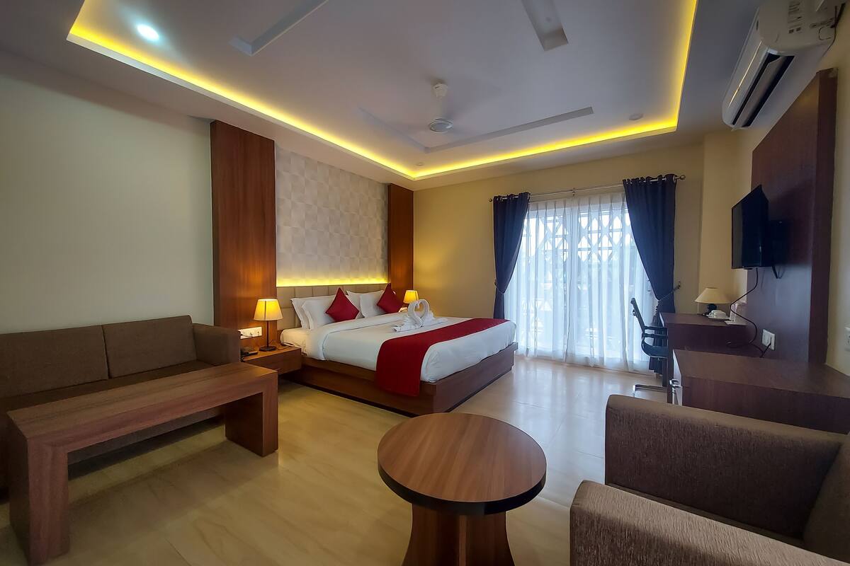 A hotel room with a bed, a sofa, a small table, a TV, and an air conditioning unit.