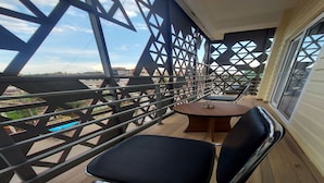 Un balcón con barandilla de celosía metálica, una mesa redonda con una silla y vistas de la ciudad.