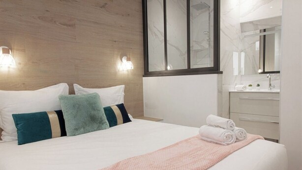 Ett hotellrum med en sänggavel i trä, en stor säng med vita sängkläder, turkosa och svarta kuddar, en rosa stickad pläd och en prydligt bäddad säng med hoprullade handdukar.