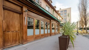 Un bâtiment doté de grandes portes et fenêtres en bois, d’un auvent vert et d’un bac à fleurs orné de plantes, placé sur le trottoir.