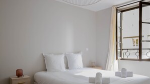 Une chambre à coucher au style minimaliste, avec un grand lit, du linge de lit blanc, une table de chevet avec une lampe et une vue sur l’extérieur par une fenêtre ouverte.