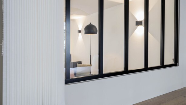 Un intérieur moderne avec une grande fenêtre, un lampadaire et un lit visible à travers la fenêtre.