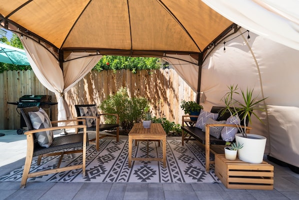 A covered outdoor seating area with a table, benches, and potted plants.