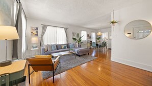 Ein modernes Wohnzimmer mit einer Couch, einem Couchtisch und Sesseln. Es gibt einen Essbereich mit Tisch und Stühlen. Der Raum hat Holzboden und weiße Wände.