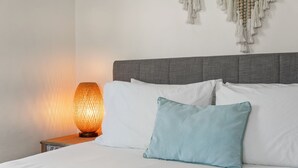 Ein ordentlich bezogenes Bett mit weißen und hellblauen Kissen, einem strukturierten Wandbehang und einer Lampe auf einem hölzernen Nachttisch.