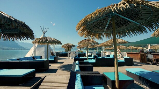 Eine Terrasse mit strohgedeckten Sonnenschirmen, Liegestühlen und einem weißen Gebäude im Hintergrund.