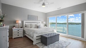 Ein Schlafzimmer mit großem Fenster, einem Bett, einem Nachttisch, einer Lampe, einem Deckenventilator und Blick auf das Wasser.