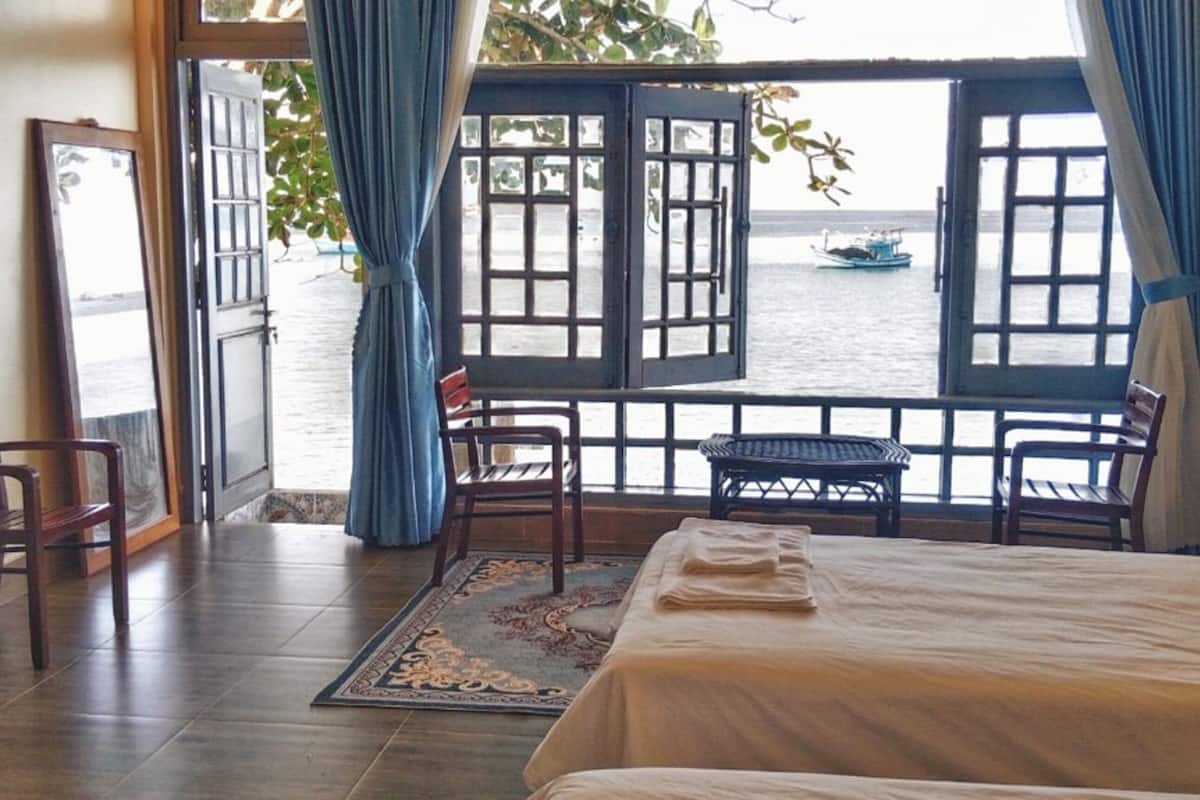 A room with a bed, a desk, and a view of the sea through open windows.