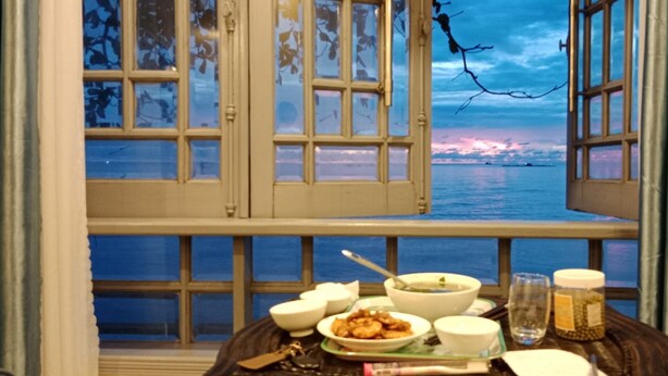 Ett matbord med tallrikar och ett glas vatten, placerat med utsikt över havet och en solnedgång som syns genom ett öppet fönster.