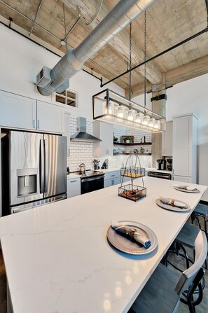 Une cuisine moderne dotée d’un grand îlot central, d’appareils électroménagers en acier inoxydable et de luminaires suspendus.