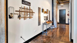 A hallway with a polished concrete floor, a wooden ceiling, and a wall-mounted sign that reads "Share Your Waco Favorites!" There is a metal rack with a lantern and a small table with a lamp and a framed picture. - The Crossing at 202- Downtown Waco/ Steps to Silos (Waco)
