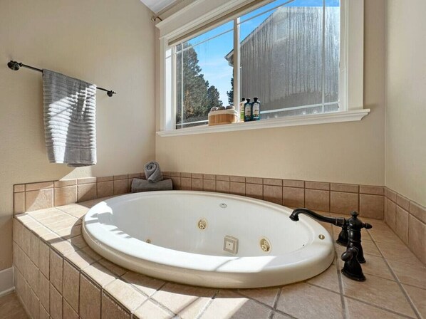 Un baño con una bañera ovalada grande, una ventana con vista a un edificio de madera y un estante con artículos de aseo.