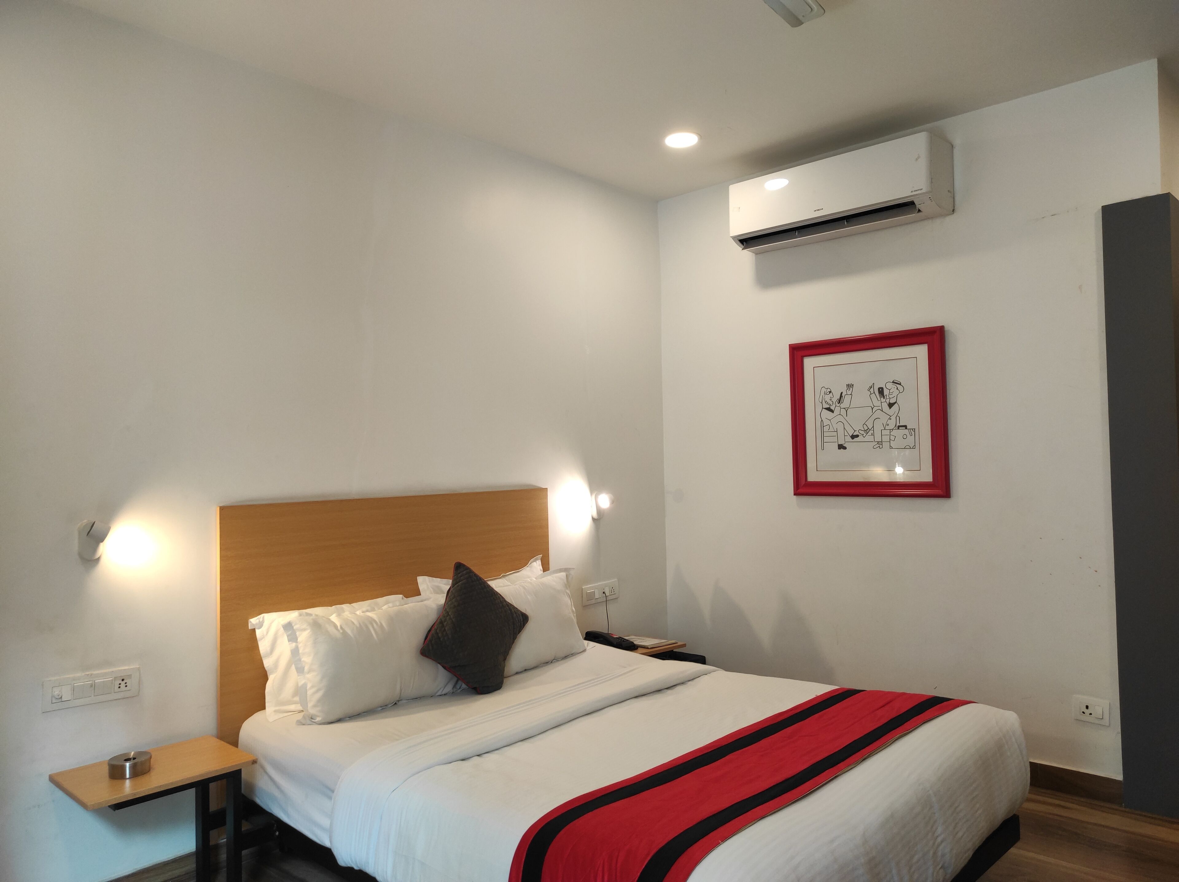 Una cama bien hecha con ropa blanca y una colcha a rayas negra y roja, un cabecero de madera, una mesita de noche con una lámpara y un equipo de aire acondicionado en la pared.