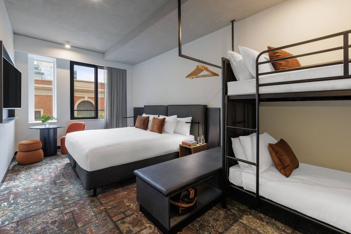 A hotel room with two beds, a bench, a TV, and a window with a view of a building.