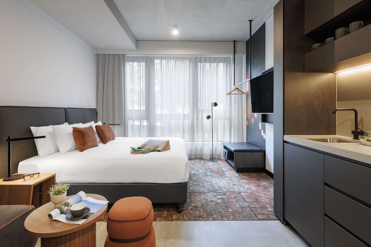 A modern hotel room with a large bed, a desk with a chair, a small table with a plant and a cup, a bench, a large window with sheer curtains, a modern kitchen area with a sink and countertop, and a television mounted on the wall.