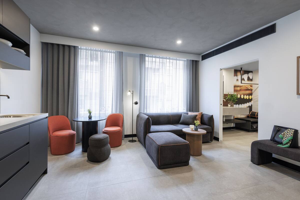 A modern living room with a sofa, ottoman, and chairs, a kitchen area with cabinets, and large windows with curtains.