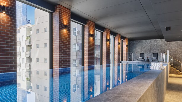 A modern indoor swimming pool with clear glass walls, a brick wall on one side, and a concrete edge. - Veriu Queen Victoria Market (Melbourne)