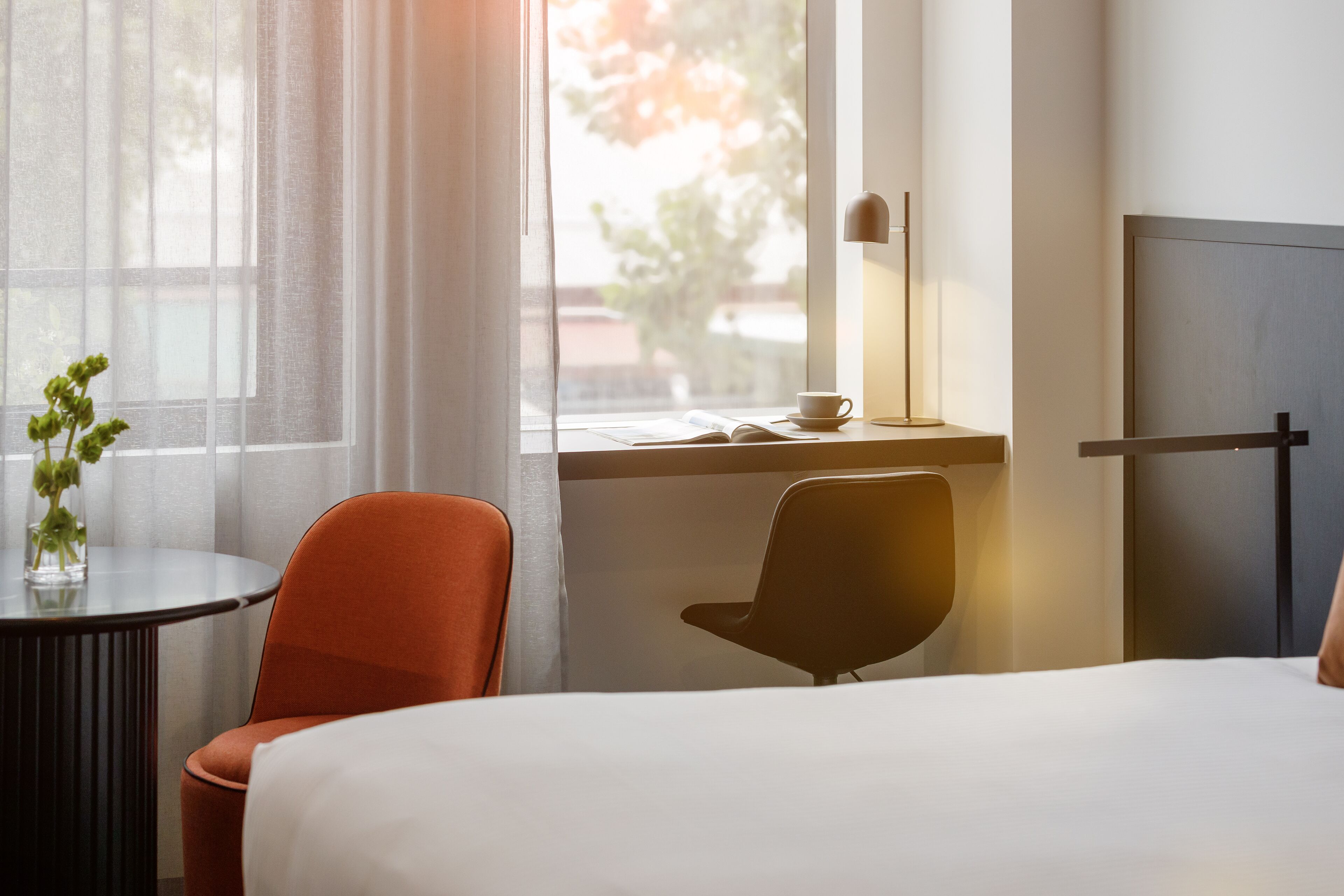 A hotel room with a bed, a desk with a cup and book, a chair, and a window with curtains.