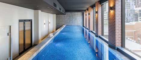 A long indoor swimming pool with blue tiles, a glass wall on one side, and a brick wall on the other.
