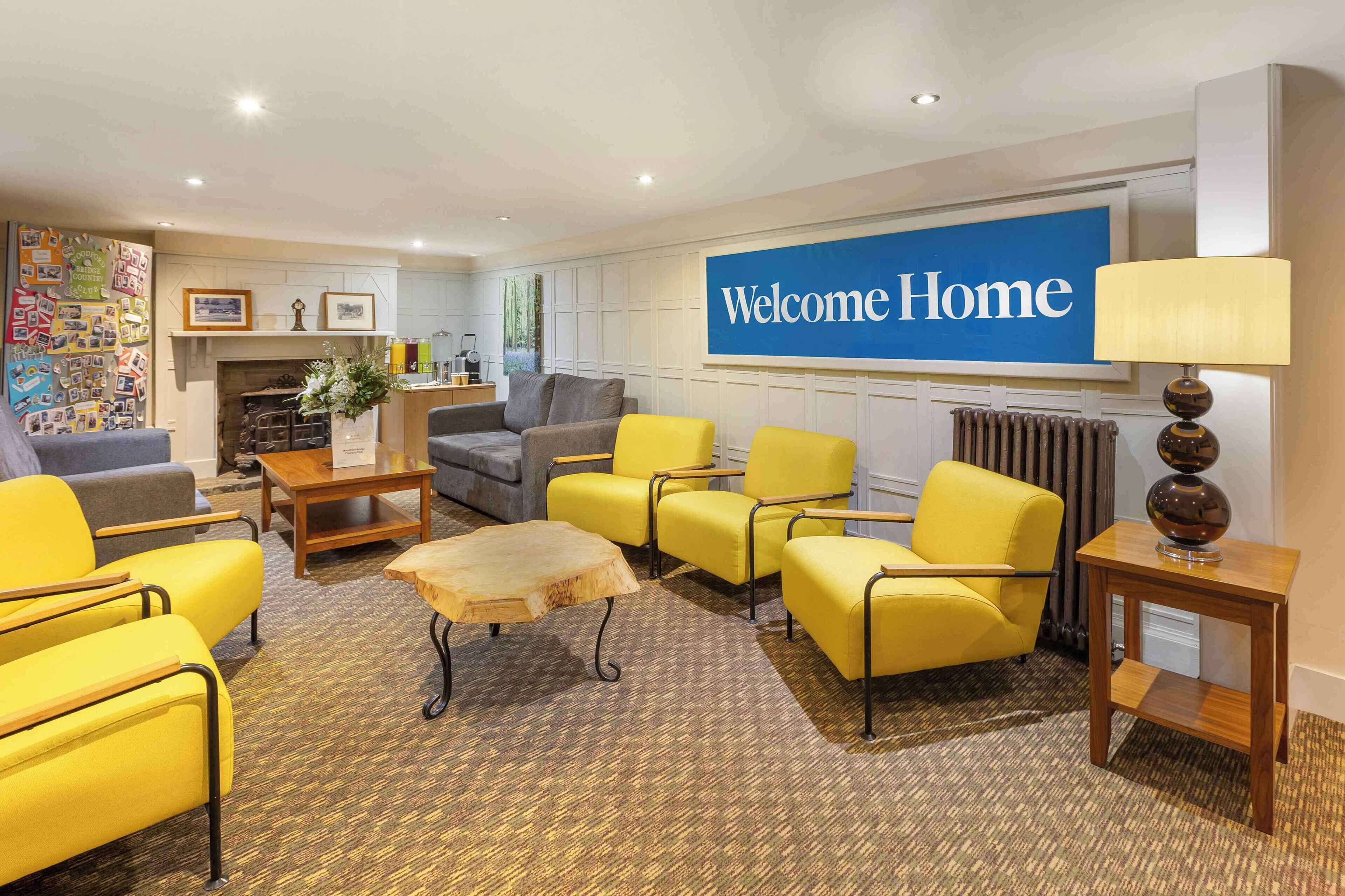 A hotel lobby with a 'Welcome Home' sign, yellow armchairs, a grey sofa, a wooden coffee table, and a fireplace.