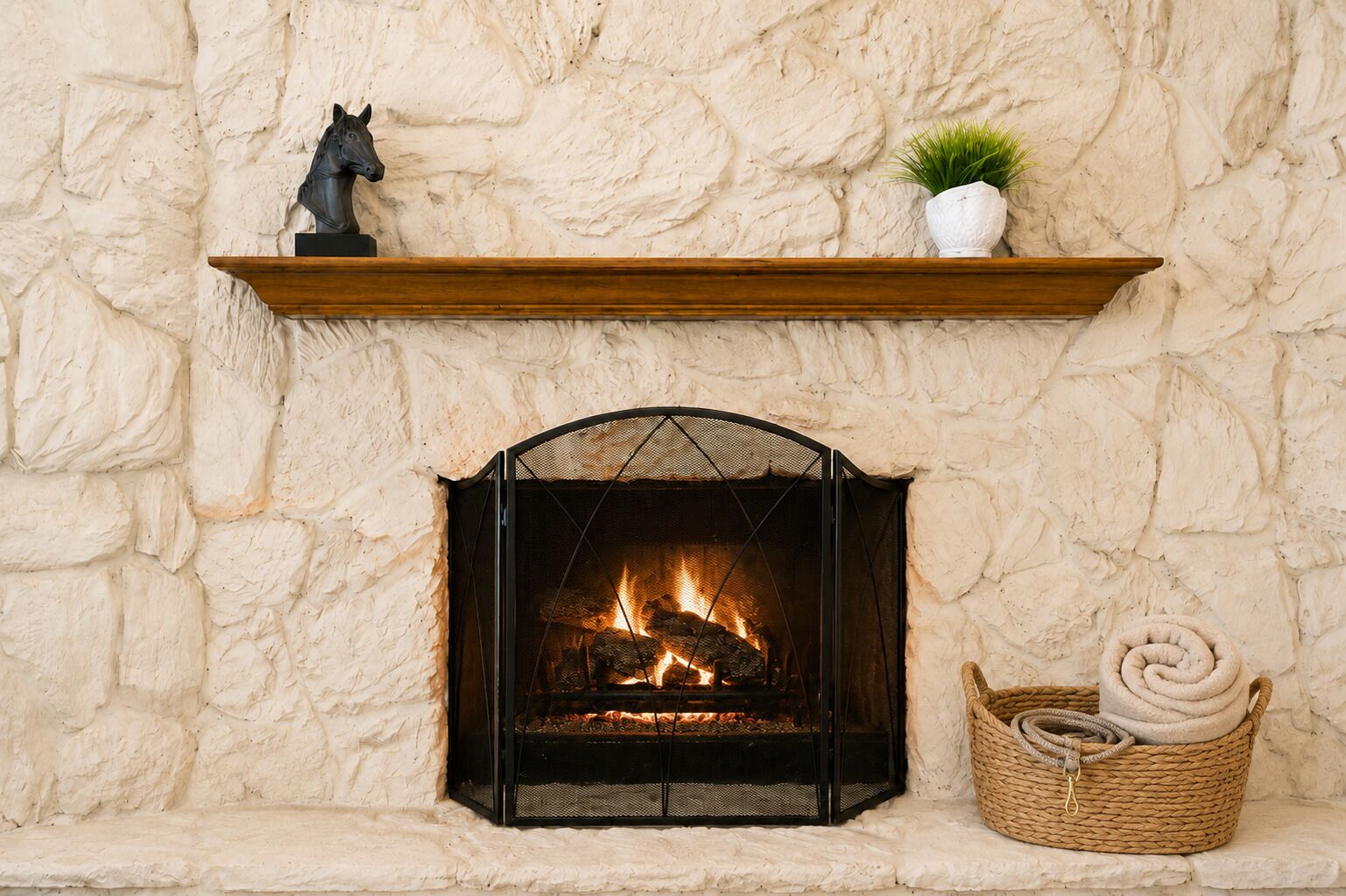 A fireplace with a stone wall, a shelf with a horse statue and a plant, and a basket with a rolled-up item.