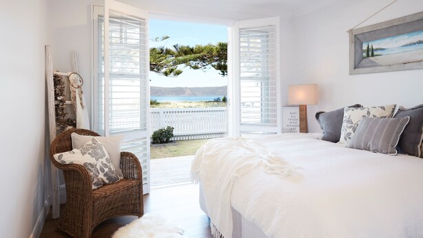 A bedroom with a large bed, a wicker chair with a pillow, a small white rug, and a view of the sea through open shutters. - Beach Front House Outdoor Hot Tub Sauna Stunning Views (Patonga)