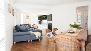 A modern living room with a blue sofa, wicker chairs, a wooden dining table, and a TV mounted on the wall. - Beach Front House Outdoor Hot Tub Sauna Stunning Views (Patonga)