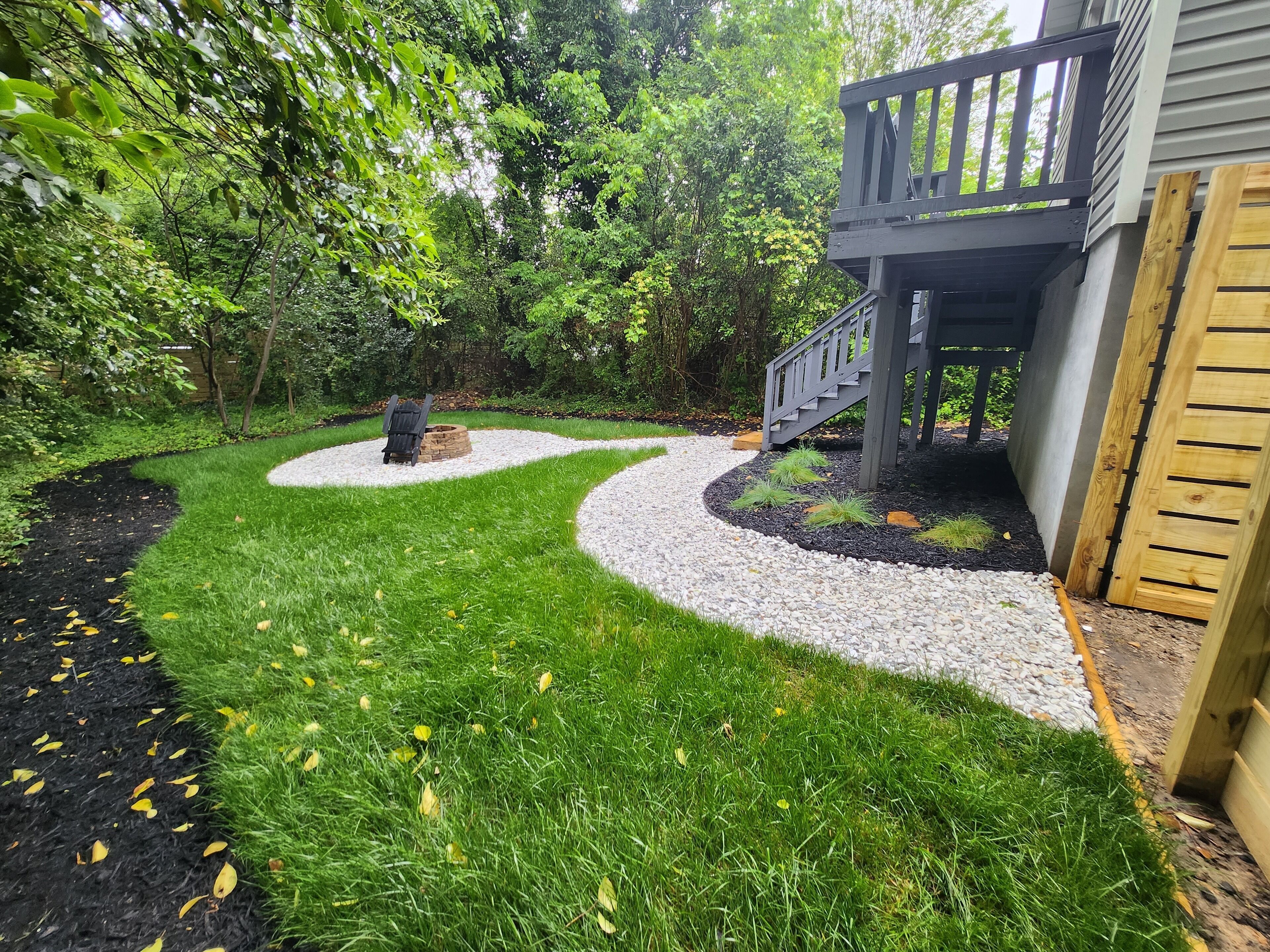 Un jardin aménagé avec un sentier sinueux recouvert de gravier, une terrasse en bois et un escalier menant à un bâtiment.
