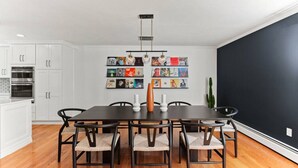 A modern kitchen with white cabinets, a dark wooden dining table, and six chairs. There is a wall-mounted light fixture above the table and a row of framed pictures on the wall.