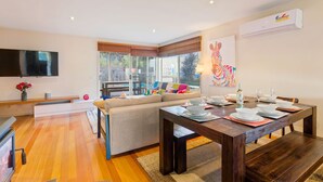 Un salon moderne avec une table à manger en bois, préparée pour quatre personnes, un grand téléviseur à écran plat, un canapé agrémenté de coussins colorés et une grande fenêtre équipée de stores.